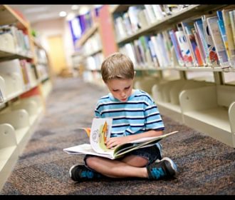 kid reading book on floor in library between rows of library shelves