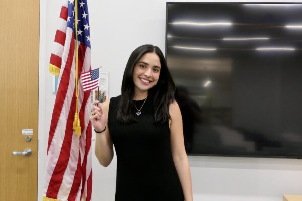 photograph of a woman in a black dress with long black hair smiling and holding a miniature us flag and standing in front of a larger us flag