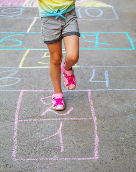photograph of a child jumping a hopscotch only the legs of the child are shown - they are wearing pink shoes and grey shorts, the hopscotch is in a variety of colors of chalk - the child is on the number 2