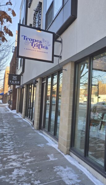 Exterior of Tropes & Trifles, a romance bookshop in Minneapolis, with a hanging white sign reading “Tropes & Trifles: A Romance Bookshop” in purple lettering above a snowy sidewalk and large street-facing windows.