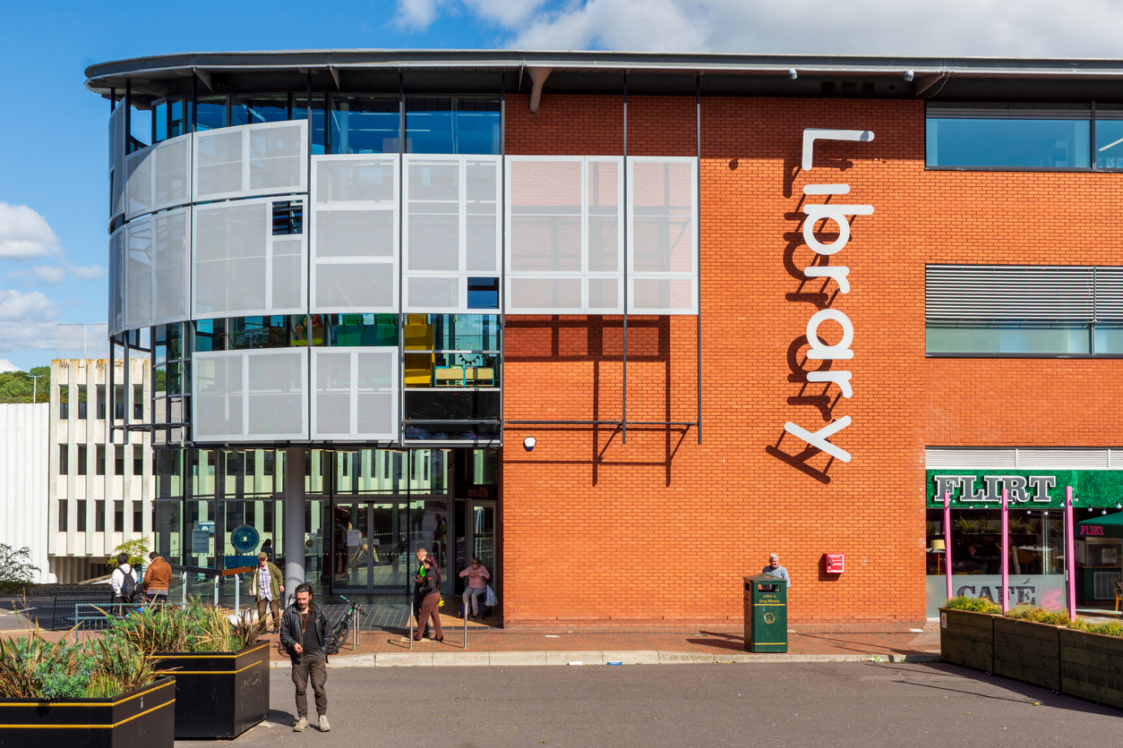 Exterior of a modern library building with a curved glass facade and an orange brick wall featuring large vertical lettering that reads “Library.” Several people are walking or sitting outside near the entrance on a sunny day.
