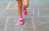 photograph of a child jumping a hopscotch only the legs of the child are shown - they are wearing pink shoes and grey shorts, the hopscotch is in a variety of colors of chalk - the child is on the number 2